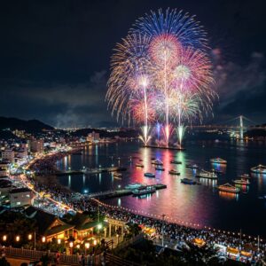 Colorful fireworks exploding over harbor with boats and a large crowd along the waterfront at night